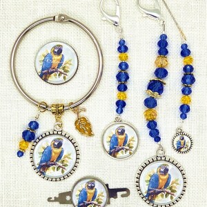 May include: A collection of craft accessories featuring a blue and yellow macaw illustration. Items include a circular ring, stitch markers with blue and yellow beads, and a key-shaped tool. The accessories have silver-colored metal accents.