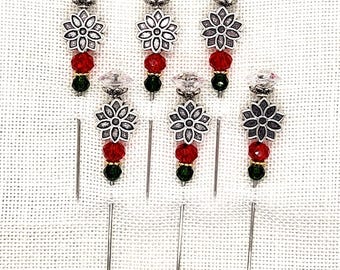 May include: Six decorative pins with silver-colored floral designs, red and green beads, and clear crystal tops. The pins are arranged vertically against a textured white background, showcasing their elegant details and festive color scheme.