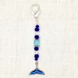 May include: A beaded charm with a silver clasp and a blue whale tail pendant. The charm features a string of blue and clear beads, with a light blue bead in the center. The whale tail is a vibrant blue color.