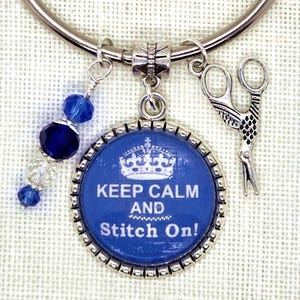 Puede incluir: Una pulsera de abalorios plateada con elementos azules y plateados. Presenta un colgante redondo azul con la inscripci&oacute;n "KEEP CALM AND Stitch On!" y una corona. Otros abalorios incluyen unas tijeras y cuentas azules.