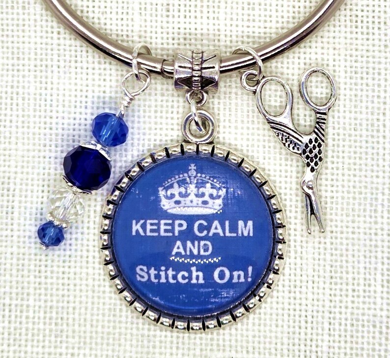 Puede incluir: Una pulsera de abalorios plateada con elementos azules y plateados. Presenta un colgante redondo azul con la inscripci&oacute;n "KEEP CALM AND Stitch On!" y una corona. Otros abalorios incluyen unas tijeras y cuentas azules.