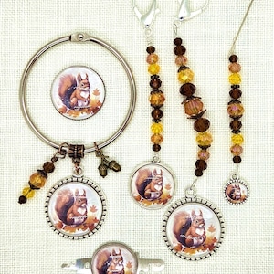 May include: A collection of handmade jewelry and accessories featuring a squirrel design. Includes a circular pendant, stitch markers, and a pin, all with a silver-tone finish and autumn-themed accents. The squirrel is depicted sitting among fall leaves.