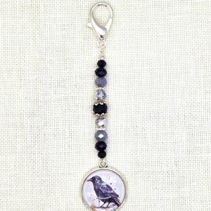 May include: A silver-toned charm with a raven illustration. The charm features a series of black, gray, and purple beads, and a silver clasp. The raven is depicted in a watercolor style, with a black and gray color palette.