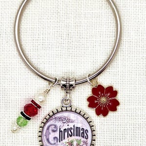 May include: A silver-colored metal ring with Christmas-themed charms. Charms include a red flower, a round charm with the word "Christmas", and a strand of red, green, and white beads. The ring is designed for decorative purposes.