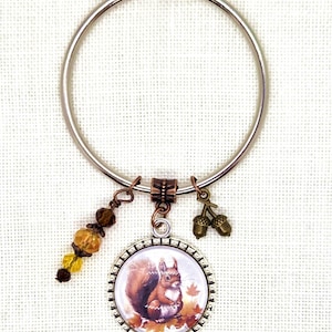 May include: A silver-tone keyring with a pendant featuring a detailed illustration of a squirrel surrounded by autumn leaves. The keyring also includes acorn charms and a strand of amber-colored beads, creating a fall-themed accessory.