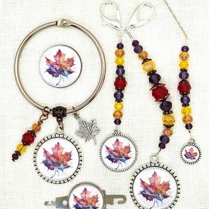 May include: A collection of fall-themed craft accessories. Includes a large silver ring, stitch markers, and a needle, all adorned with a watercolor maple leaf design in shades of red, orange, and purple. The stitch markers feature colorful beaded accents.