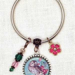 May include: A silver metal keyring with decorative charms. The charms include a round pendant with an owl illustration, a pink flower, and a strand of beads in pink, green, and clear. The owl pendant has a blue background.