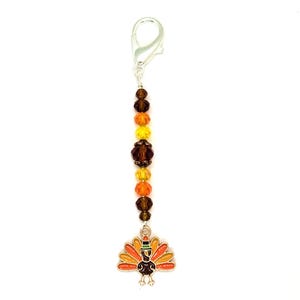 May include: A silver-toned lobster clasp keychain featuring a vertical strand of faceted beads in shades of brown, orange, and yellow. At the base hangs a metal charm shaped like a turkey with colorful enamel detailing in orange, yellow, and brown.