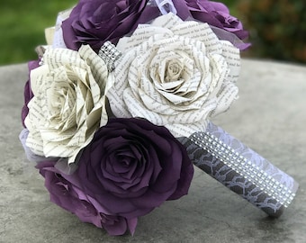 May include: A bouquet of handcrafted paper flowers featuring deep purple solid-colored roses and cream-colored roses made from printed book pages. The handle is wrapped in grey lace fabric and accented with a band of sparkling silver rhinestones.