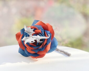 May include: A handcrafted paper rose with alternating layers of navy blue and rust-colored petals. A silver-colored dragon figurine is perched on top. The stem is a muted gray color. The background is blurred.