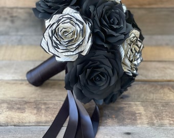 May include: A bouquet of handcrafted paper flowers featuring solid black roses interspersed with roses made from printed book pages. The stems are wrapped in a dark ribbon with long, flowing black ribbon tails extending from the base of the bouquet.