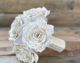 May include: A bouquet of white paper roses made from the pages of a book. The roses are arranged in a circular shape and are tied together with a white ribbon. The bouquet is sitting on a wooden surface.