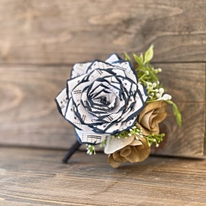 May include: A handcrafted paper flower boutonniere featuring a large white rose made from sheet music with navy blue edges. Accompanying the rose are smaller brown paper flowers and sprigs of greenery. The stem is black.
