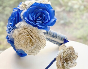 May include: A blue and white paper flower bouquet with a smaller boutonniere. The bouquet is made of paper roses with blue petals and white paper roses made from book pages. The boutonniere is also made of paper roses with a blue ribbon.