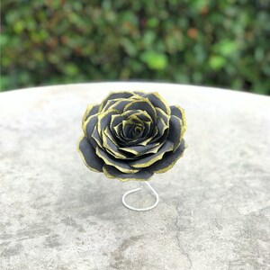 May include: A handcrafted paper rose in black with gold glitter edges, displayed on a white wire stand. The flower is set against a blurred green background, showcasing the intricate details of the petals.