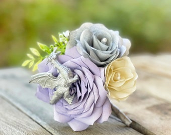 May include: A bouquet of artificial flowers with a silver dragon charm. The bouquet includes a lavender rose, a cream rose, and a silver-grey rose.
