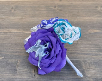 May include: A purple paper rose with a silver dragon figurine in the center. A teal paper rose with text from a book is behind the purple rose. The roses are attached to a white stem.