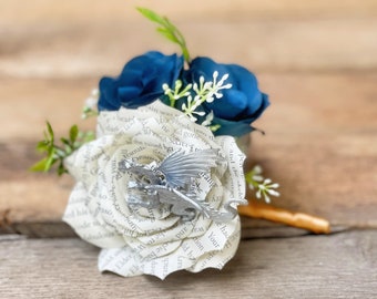 May include: A white paper rose with a silver dragon charm, surrounded by two blue fabric roses and white sprigs. The rose is made from the pages of a book.