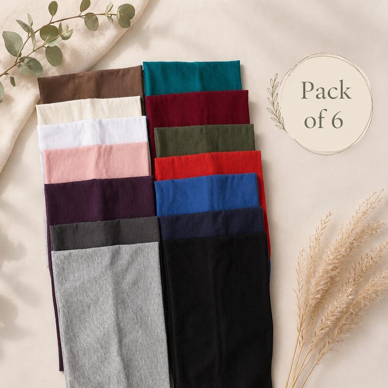 May include: A pack of six headbands in various colours, including brown, cream, white, pink, purple, grey, teal, burgundy, olive green, red, blue, and black. The headbands are neatly arranged on a neutral-toned surface.