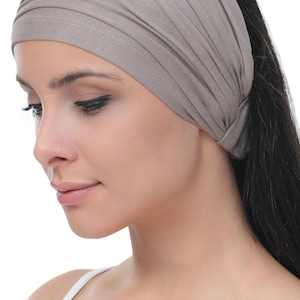 May include: A wide, taupe-coloured headband made from soft fabric. The headband is gathered at the top and worn across the forehead. The model is wearing a white vest. The background is white.