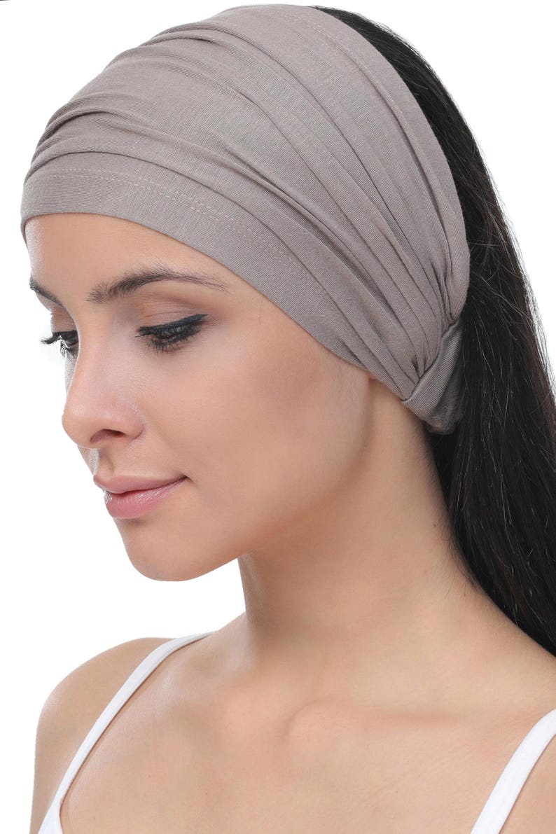 May include: A wide, taupe-coloured headband made from soft fabric. The headband is gathered at the top and worn across the forehead. The model is wearing a white vest. The background is white.