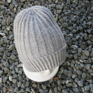 May include: A grey knitted beanie hat with a ribbed texture. The hat is sitting on a grey and white surface.