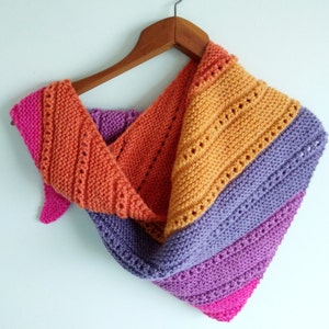 Puede incluir: Un chal triangular de punto en tonos de rosa, morado, naranja y amarillo. El chal está colgado en una percha de madera.
