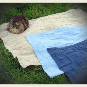 May include: A fluffy cat rests on a beige knitted blanket with a geometric pattern. Below, two additional blankets in light blue and navy blue are visible, also with the same pattern. The blankets are displayed on green grass.
