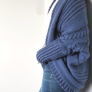 May include: A blue knitted cardigan with a cable knit pattern on the sleeves. The cardigan is open at the front and has a relaxed fit.