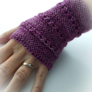 Op de afbeelding: Een gebreide polswarmers in een diepe paarse tint. De manchet heeft een textuurpatroon met kleine openingen. Het item wordt op een hand gedragen, met een gouden ring zichtbaar op de ringvinger. De achtergrond is een effen wit oppervlak.