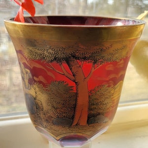 May include: A red glass goblet with a gold rim and base. The goblet is decorated with a gold painted scene of a tree and foliage.