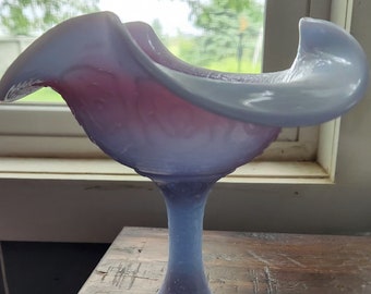 May include: A light blue and pink glass bowl with a ruffled edge and a pedestal base. The bowl is made of iridescent glass and has a vintage look.