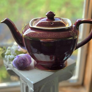 May include: A deep burgundy teapot with a gold crackle glaze finish and gold trim along the lid, spout, and handle. The teapot features a square-shaped lid with a small knob and a wide, rounded body that tapers down to a pedestal base.