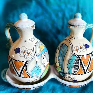 May include: Two ceramic oil and vinegar cruets with matching tray. The cruets are off-white with blue, orange, and green accents. Each cruet has a handle and a lid. One cruet says "ACE" and the other says "OLIO". The tray is off-white with a blue trim.