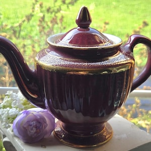 May include: A deep burgundy ceramic teapot featuring a ribbed body, a matching lid with a pointed finial, and gold-colored trim along the rim, base, and spout. The teapot has a curved handle and a pedestal base, with a glossy finish throughout.