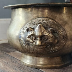 May include: A large, antique brass bowl with an ornate design featuring a stylized animal face. The bowl has a wide, rounded base and a flared rim.