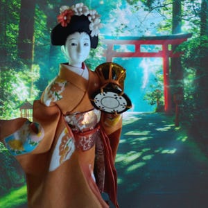May include: A Japanese geisha doll wearing a traditional orange kimono with floral accents and a black wig adorned with flowers. The doll holds a decorative fan and stands in front of a scenic background featuring a red torii gate and a forest path.