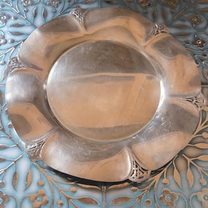 May include: A silver-colored, decorative plate with a scalloped edge and ornate detailing. The plate has a circular center and a slightly raised rim. The design includes decorative flourishes at the edge of each scallop. The plate is likely made of metal.