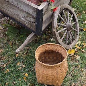 May include: A rustic wooden cart with a red flower pot and vibrant red flowers. A woven basket sits on the grass in front of the cart. The cart has a weathered appearance, with a large wooden wheel.