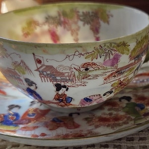 May include: A vintage teacup and saucer set with a Japanese-inspired design. The cup and saucer feature a detailed landscape scene with figures, buildings, and a Japanese flag. The set has a white base with colorful accents and a gold trim.