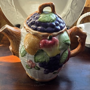 May include: A ceramic teapot with a fruit basket design. The lid is adorned with purple grapes and green leaves. The body features apples, pears, and more grapes. The handle and spout are brown. The teapot is on a dark wooden surface.