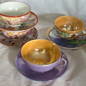 May include: Several antique teacups and saucers. Two sets of teacups and saucers have a detailed, colorful design with figures and landscapes. One teacup has a gold interior and a purple saucer. Another teacup has a gold interior with a landscape design.