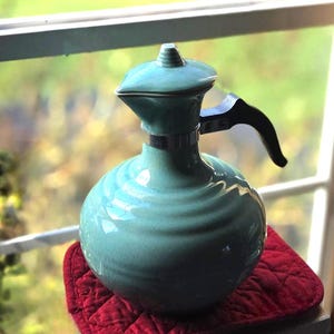 May include: A turquoise ceramic carafe with a rounded, ribbed body and a glossy crackle glaze finish. The vessel features a matching lid with a pointed finial, a silver-toned metal collar, and a black handle extending from the side of the neck.