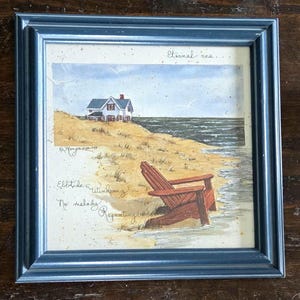 May include: A framed watercolor painting of a coastal scene. The artwork depicts a beach with a house on a dune, an Adirondack chair, and the ocean. The frame is a muted blue. Text on the painting reads "Eternal sea..." and "Ebb tide & windsong..."