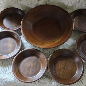 May include: A collection of wooden bowls and plates in varying sizes. The large, round bowl is surrounded by six smaller, shallow bowls. The wood grain is visible, and the color is a warm, medium brown. The items are arranged on a white, textured surface.