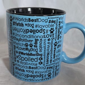 May include: A light blue ceramic mug with a black interior and handle. The mug is covered in black text and paw prints, including phrases like "World's Best Dog," "Unconditional Love," and "Spoiled." A perfect gift for dog lovers.