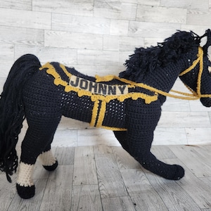 May include: Black crocheted horse with a yellow and brown saddle and bridle. The saddle has the name "JOHNNY" written on it.