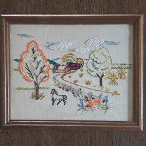 May include: Framed embroidered landscape scene featuring a horse, trees, a farmhouse, and a winding path. The artwork uses various colors of thread to depict a rural scene with a fence and flowers. The frame is made of wood.