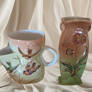 May include: A vintage ceramic mug and vase set. The mug is light green and peach with bird and gold accent designs. The vase is peach and green with a floral design and gold accents. Both pieces have a glossy finish.
