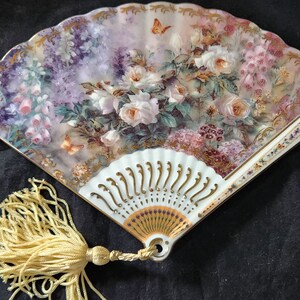 May include: A decorative hand fan featuring a floral design with white roses, pink hydrangeas, and butterflies. The fan has a white handle with gold accents and a gold tassel.